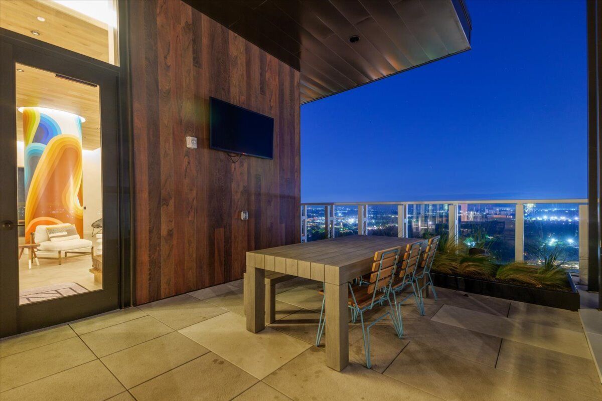 A modern outdoor dining area with a wooden table and chairs, a flat-screen TV mounted on the wall, and a view of the city at night.