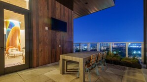 A modern outdoor dining area with a wooden table and chairs, a flat-screen TV mounted on the wall, and a view of the city at night. - King Retreat/Rooftop Views/Rainey/UT/Moody/6th St (Austin)