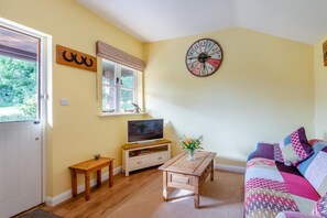 Ein Wohnzimmer mit Fernseher, Holzmöbeln, einer Uhr und einem Fenster mit Blick ins Grüne.