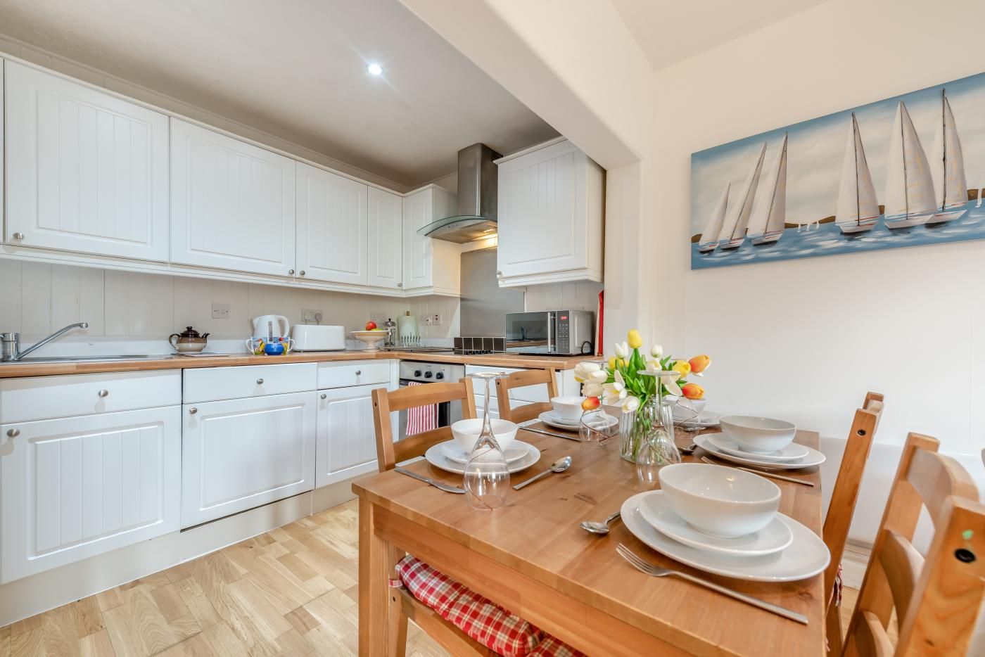 Una cocina moderna con armarios blancos, una mesa de comedor de madera dispuesta para cuatro personas y un cuadro con barcos de vela en la pared.
