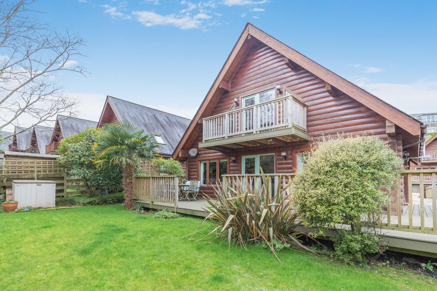 A log cabin with a balcony, surrounded by a well-maintained lawn and a wooden fence.