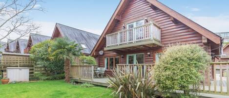 A log cabin with a balcony, surrounded by a well-maintained lawn and a wooden fence.