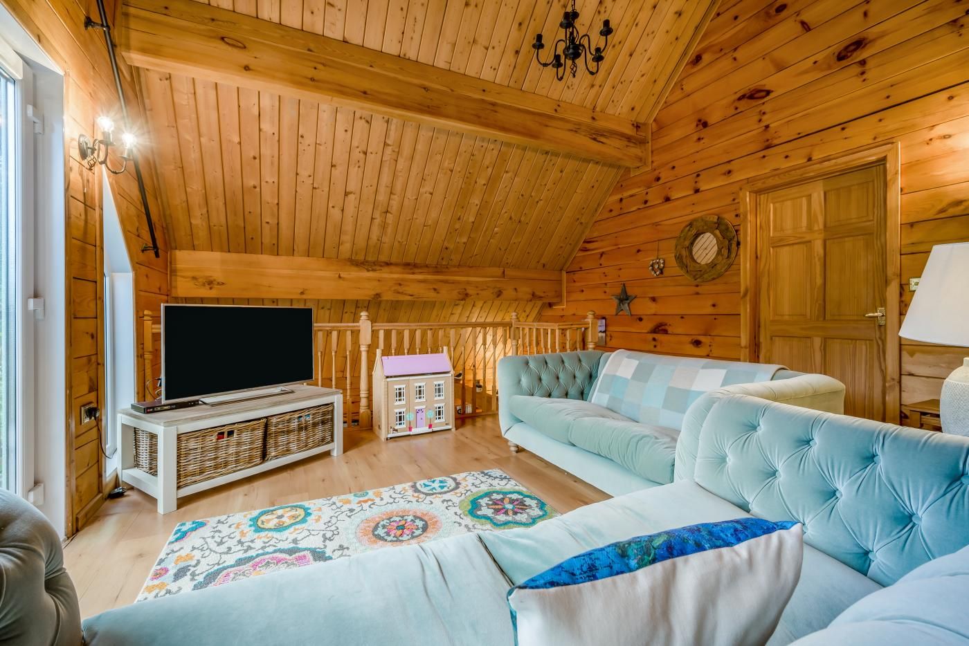 A cozy log cabin interior with a television, plush sofas, a patterned rug, and a wooden staircase.
