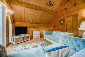 A cozy log cabin interior with a television, plush sofas, a patterned rug, and a wooden staircase.