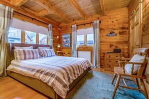 Un dormitorio tipo cabaña de madera con una cama, una silla, una mesita de noche, una lámpara y una ventana con cortinas.