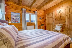 A wooden cabin bedroom with a bed, a chair, a painting, and a window. - 3 Bed in Wadebridge (oc-p34722) (Little Petherick)