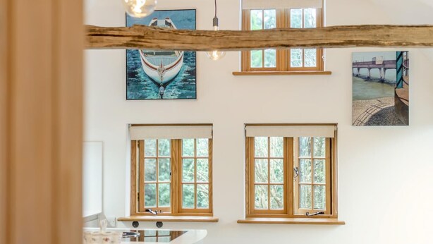 Ein Zimmer mit Holzbalkendecke, zwei Fenstern mit Holzrahmen, einer Küchenzeile mit Arbeitsplatte und Kunstwerken an den Wänden.