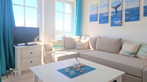 Een kamer met een televisie op een witte standaard, een beige bank, een witte bijzettafel met decoratieve objecten, en een raam met blauwe gordijnen.