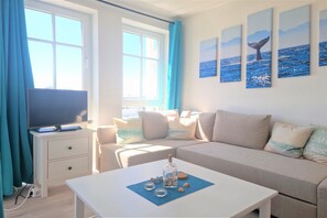 A room with a television on a white stand, a beige sofa, a white coffee table with decorative items, and a window with blue curtains. - Stilvolle Ferienwohnung mit Gemutlichem Balkon in Sellin (Sellin)