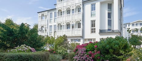Un bâtiment blanc doté de nombreux balcons et d’un jardin où poussent des fleurs roses.