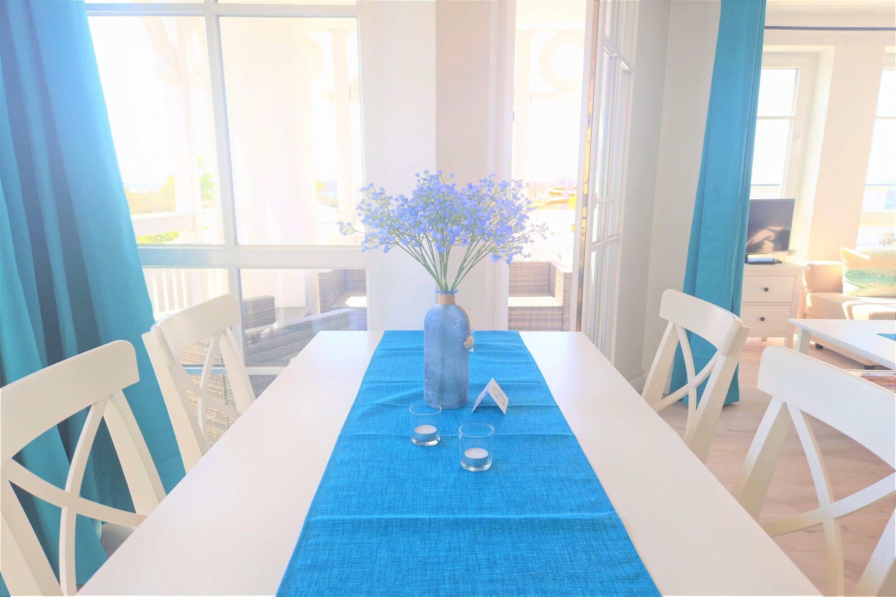 Een eettafel met een blauwe tafelkleed, witte stoelen, een vaas met bloemen en een klein bordje.