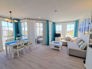 A living room with a sofa, a TV, a dining table with chairs, and a balcony with curtains. - Stilvolle Ferienwohnung mit Gemutlichem Balkon in Sellin (Sellin)
