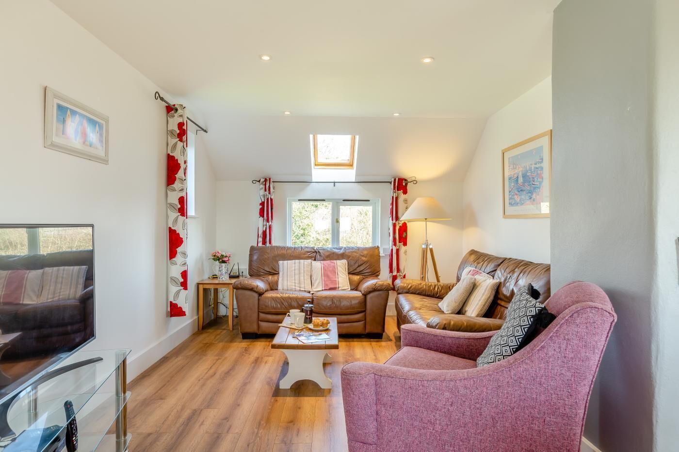Ein Wohnzimmer mit einer Couch, Sesseln, einem Couchtisch und einem Fernseher. Vorhanden sind Vorhänge, eine Lampe und ein Fenster.