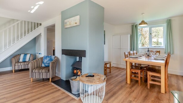 A living room with a fireplace, striped chairs, a dining table set for four, and a staircase. - 4 Bed in Marhamchurch (oc-p34749) (Marhamchurch)