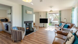 A living room with a fireplace, a sofa, a television, and a kitchen area. - 4 Bed in Marhamchurch (oc-p34749) (Marhamchurch)