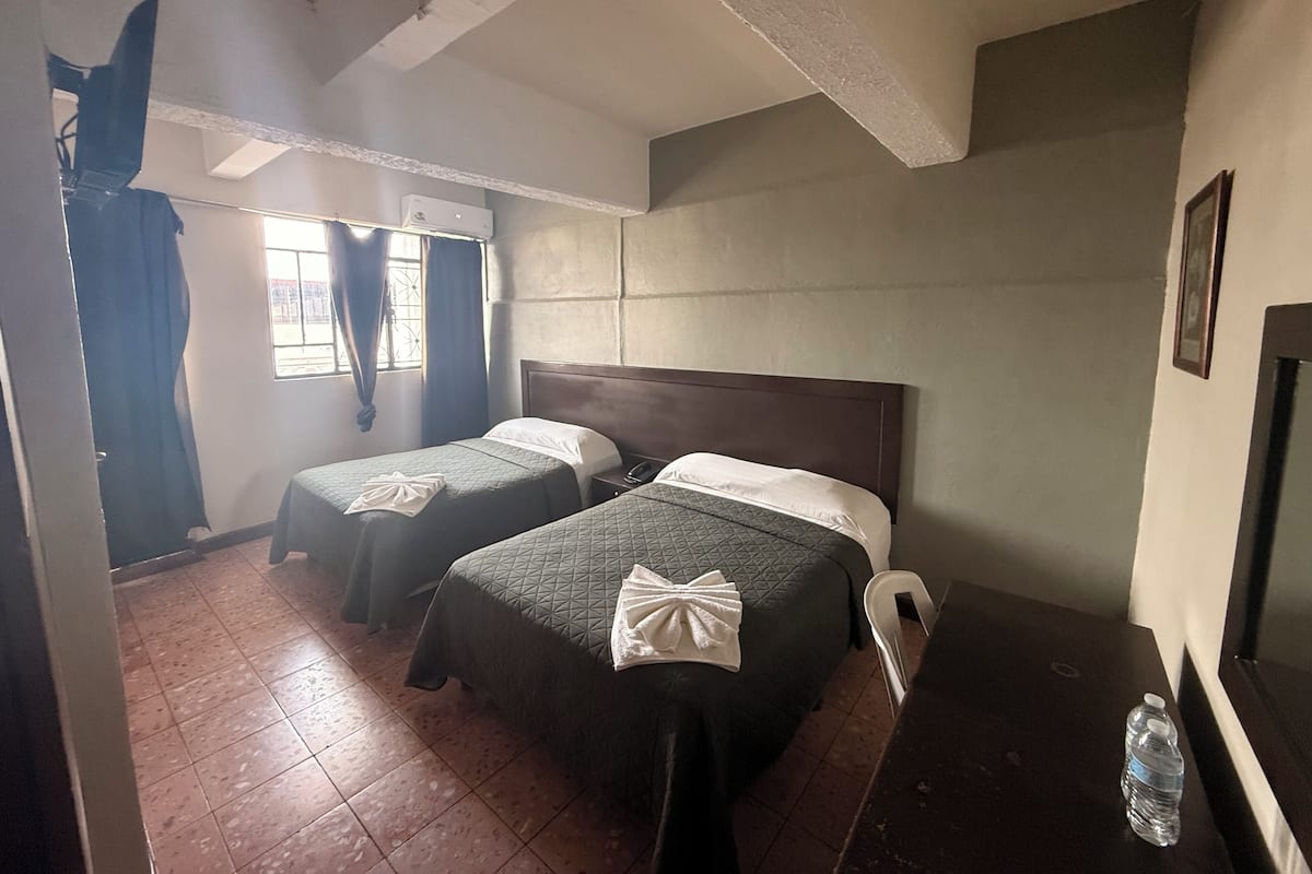 Habitación con dos camas, una ventana con cortinas, cabecera de madera, mesita de noche y una botella de agua.