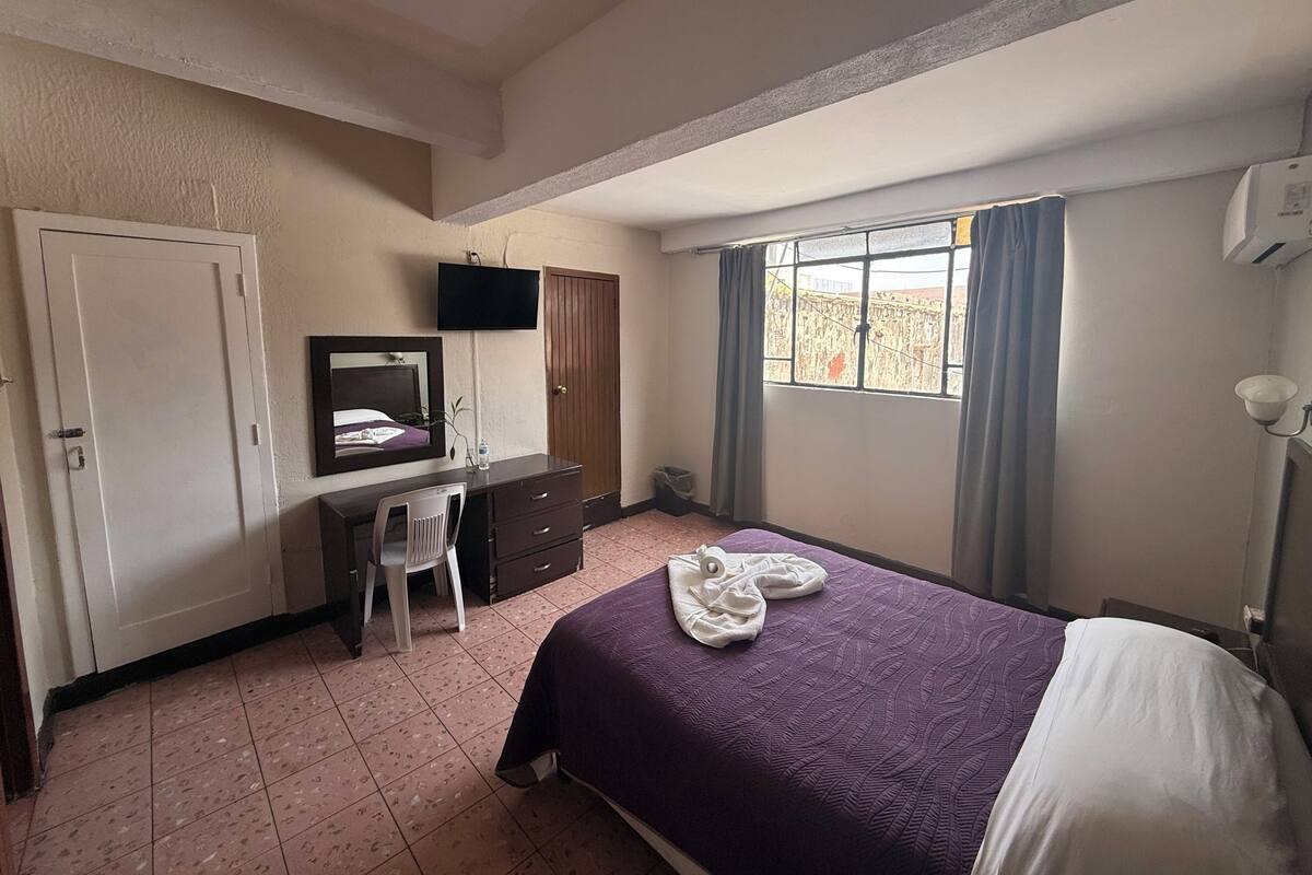 Un dormitorio con cama, escritorio, silla, televisor, armario, ventana con cortinas y aire acondicionado instalado en la pared.