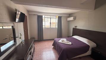 A hotel room with a bed, a TV on a dresser, a window with curtains, and a tiled floor.