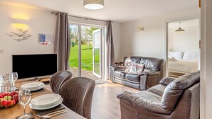 En moderne stue med en sofa, et spisebord dækket med tallerkener og glas, et fjernsyn og udsigt til en grøn have gennem en glasdør.