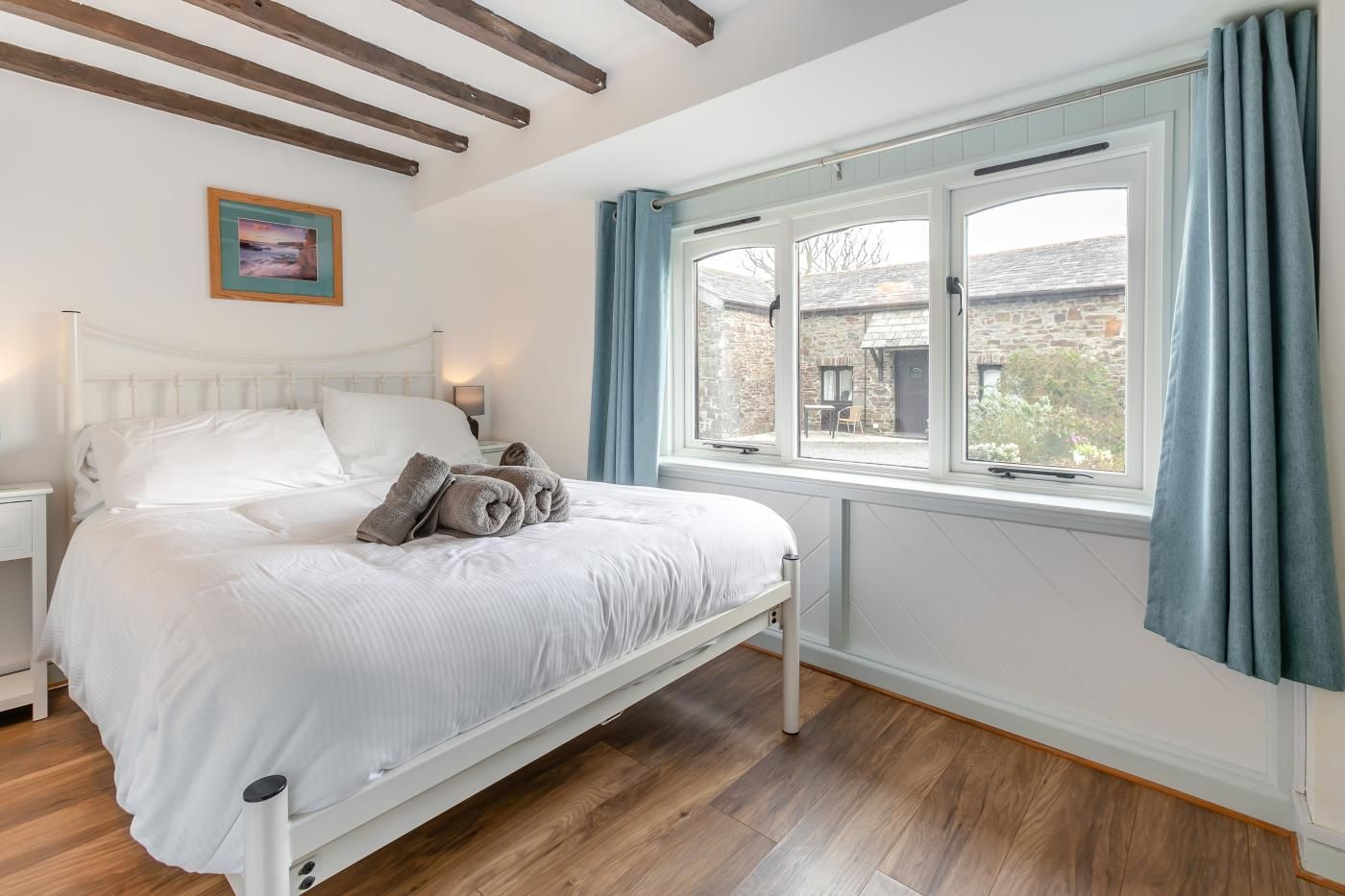 A neatly made bed with pillows and a bedside table, a window with curtains, and a painting on the wall.