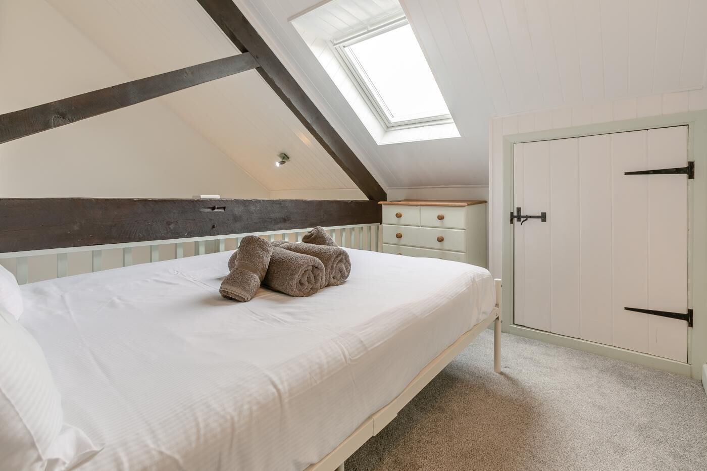 Une chambre avec un plafond en pente, un puits de lumière, un lit avec une literie blanche, une commode et une armoire.