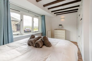A bedroom with a large bed, a dresser, and a window with a view of a car and buildings outside. - 3 Bed in Marhamchurch (oc-d34751) (Marhamchurch)
