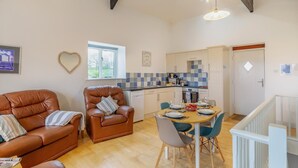 A cozy living room with a brown leather sofa, a dining table with chairs, and a kitchen area with a window. - 2 Bed in Marhamchurch (oc-s34752) (Marhamchurch)