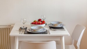 Une table à manger est dressée avec des assiettes, des bols, des couverts et des verres, et un saladier rempli de fruits est posé sur la table.