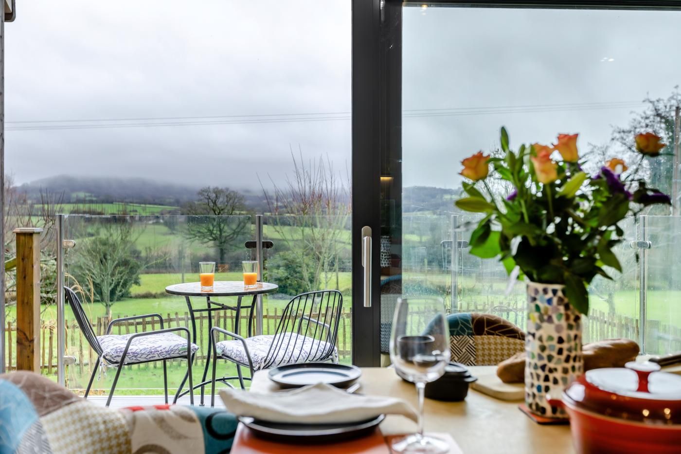 Un coin repas avec une table dressée pour deux personnes, un vase rempli de fleurs et une vue sur la campagne.