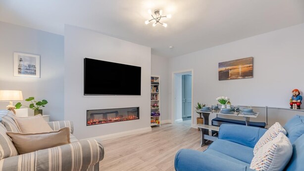 Uma sala de estar moderna com uma televisão de tela plana, lareira, estante de livros, área de jantar com mesa posta e um sofá azul.