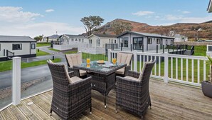 Varanda com mobiliário de vime e mesa de vidro, com vista para um parque de caravanas com edifícios brancos e uma montanha ao fundo.