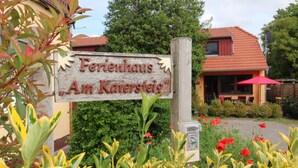 Un cartello di legno con la scritta 'Ferenhaus Am Karsteig', un edificio con tetto rosso e un giardino con fiori gialli e rossi.