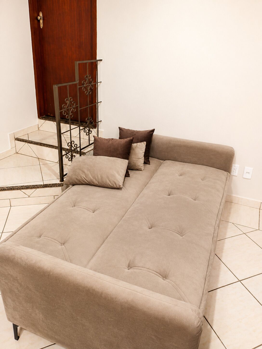 Un canapé-lit beige, doté d’une structure en métal et d’éléments décoratifs, est placé sur un sol carrelé, avec une porte en bois en arrière-plan.