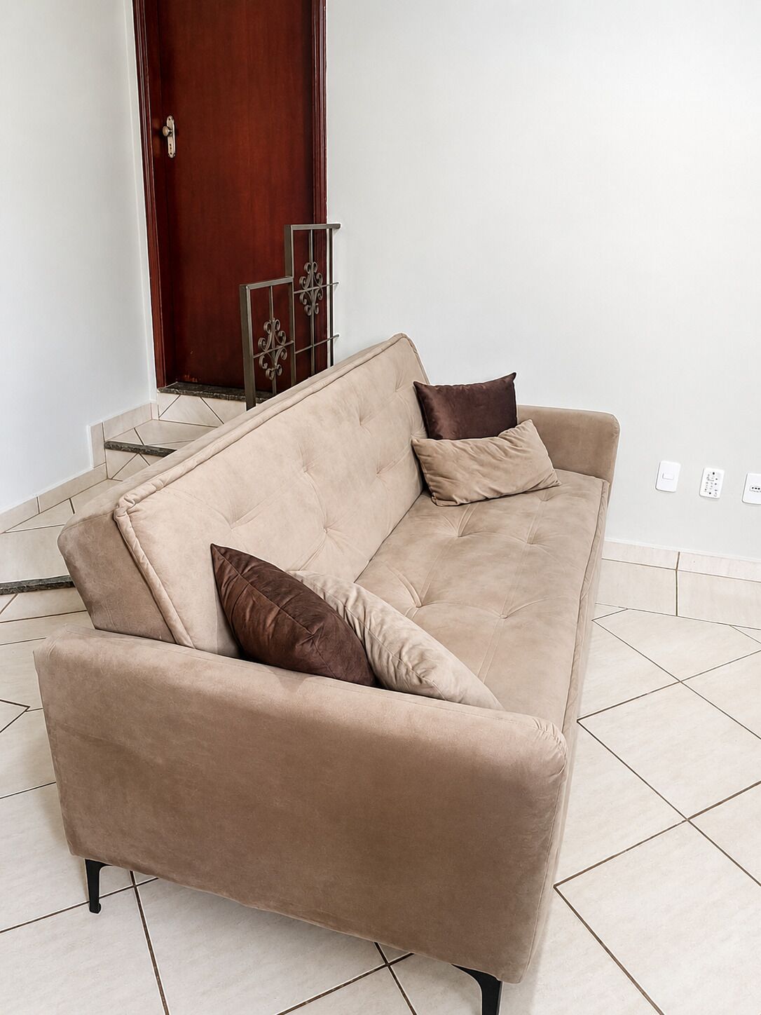 Um sofá bege com pernas pretas em um quarto com piso de azulejos e porta de madeira.