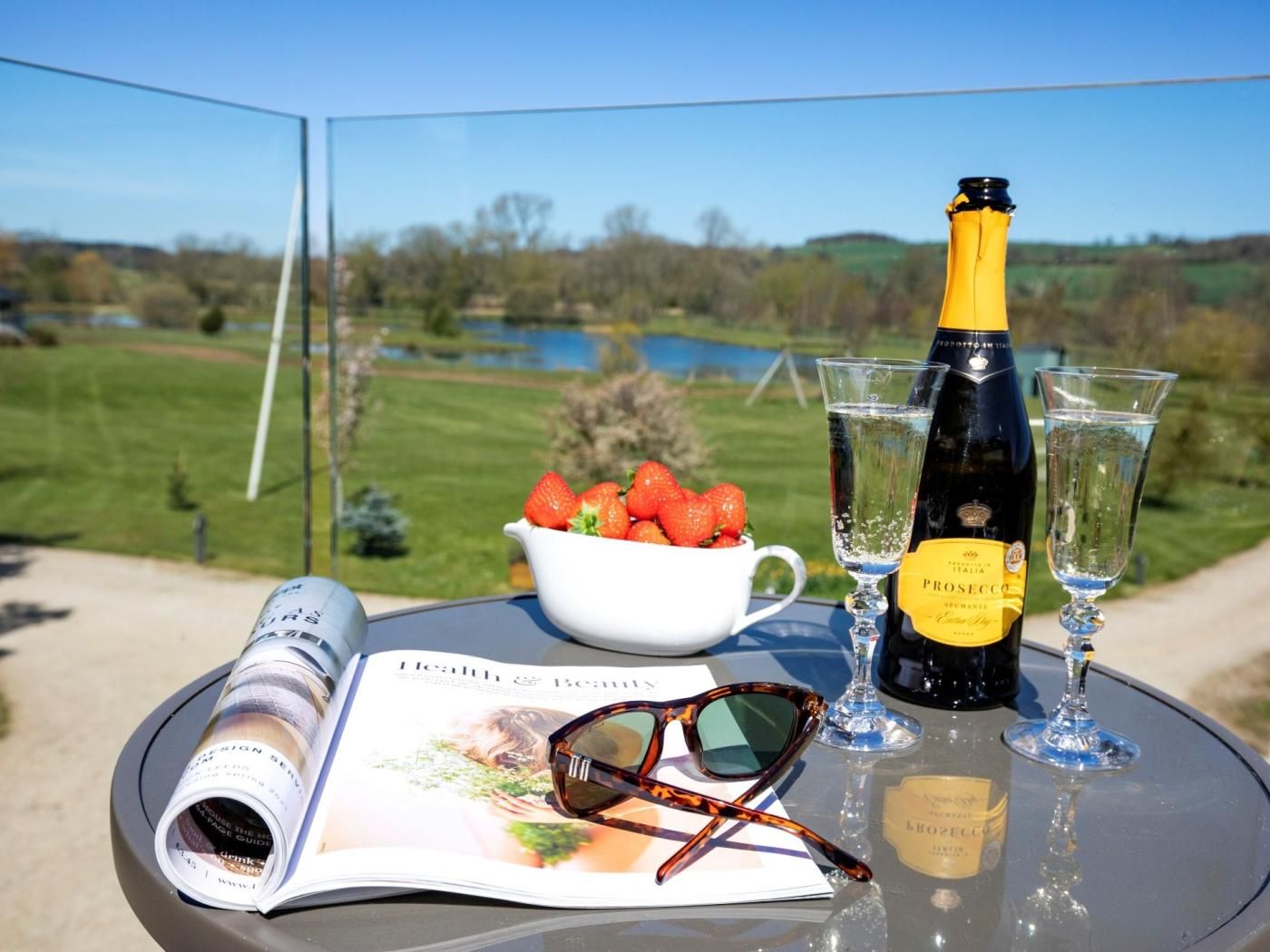A table with a bottle of prosecco, two glasses, a bowl of strawberries, a magazine, and a pair of sunglasses.