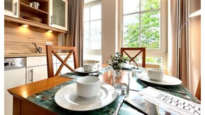 Een eetgedeelte met een houten tafel gedekt voor twee personen, een raam met uitzicht op groen en een keuken met houten kastjes en een inbouwoven.