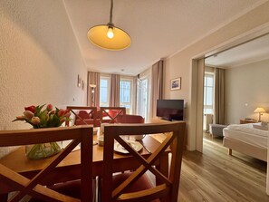 A dining area with a wooden table and chairs, a vase of flowers, a television, and a bed in the adjacent room. - Stilvoll Eingerichtete Ferienwohnung mit Grossem Balkon in Sellin (Sellin)