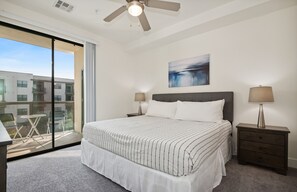 Une chambre dotée d’un grand lit, de deux tables de chevet avec des lampes, d’un ventilateur de plafond, d’une fenêtre donnant sur des immeubles et d’un balcon avec du mobilier d’extérieur.