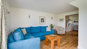 Ein Wohnzimmer mit blauem Sofa, einem Holztisch, einem beigefarbenen Sessel und einer Küche im Hintergrund.