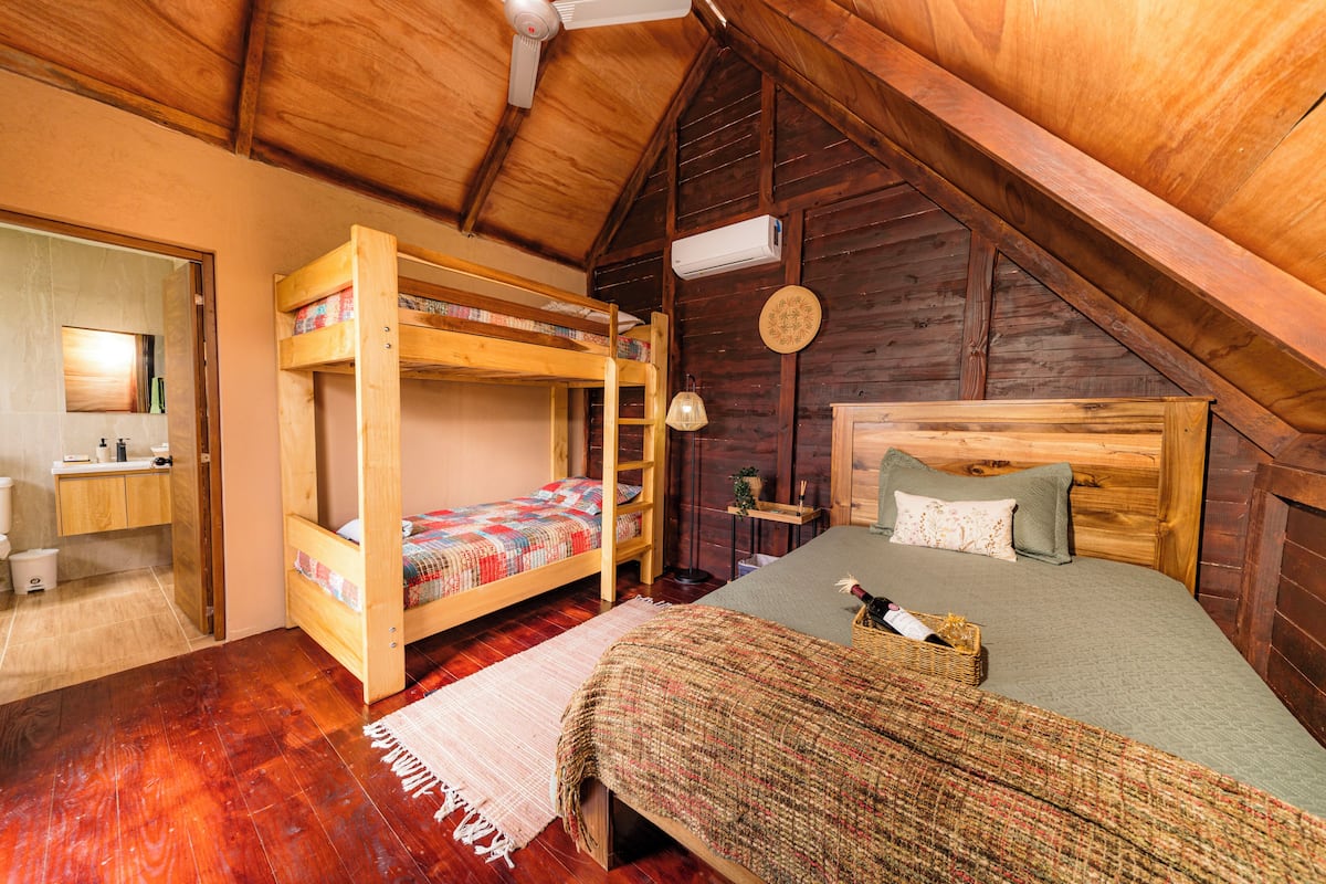 Une chambre mansardée en bois avec des lits superposés, un lit simple, un petit bureau et une fenêtre.