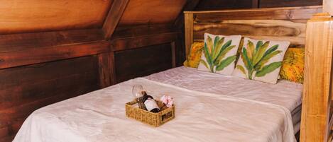 Techo de madera, cama con colcha blanca, dos almohadas con motivos florales y una cesta de mimbre con una botella y flores.