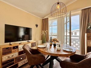 A dining area with a TV, wooden furniture, and a view of buildings through large windows. - Stilvolle Ferienwohnung mit Balkon und Wlan in Sellin (Sellin)