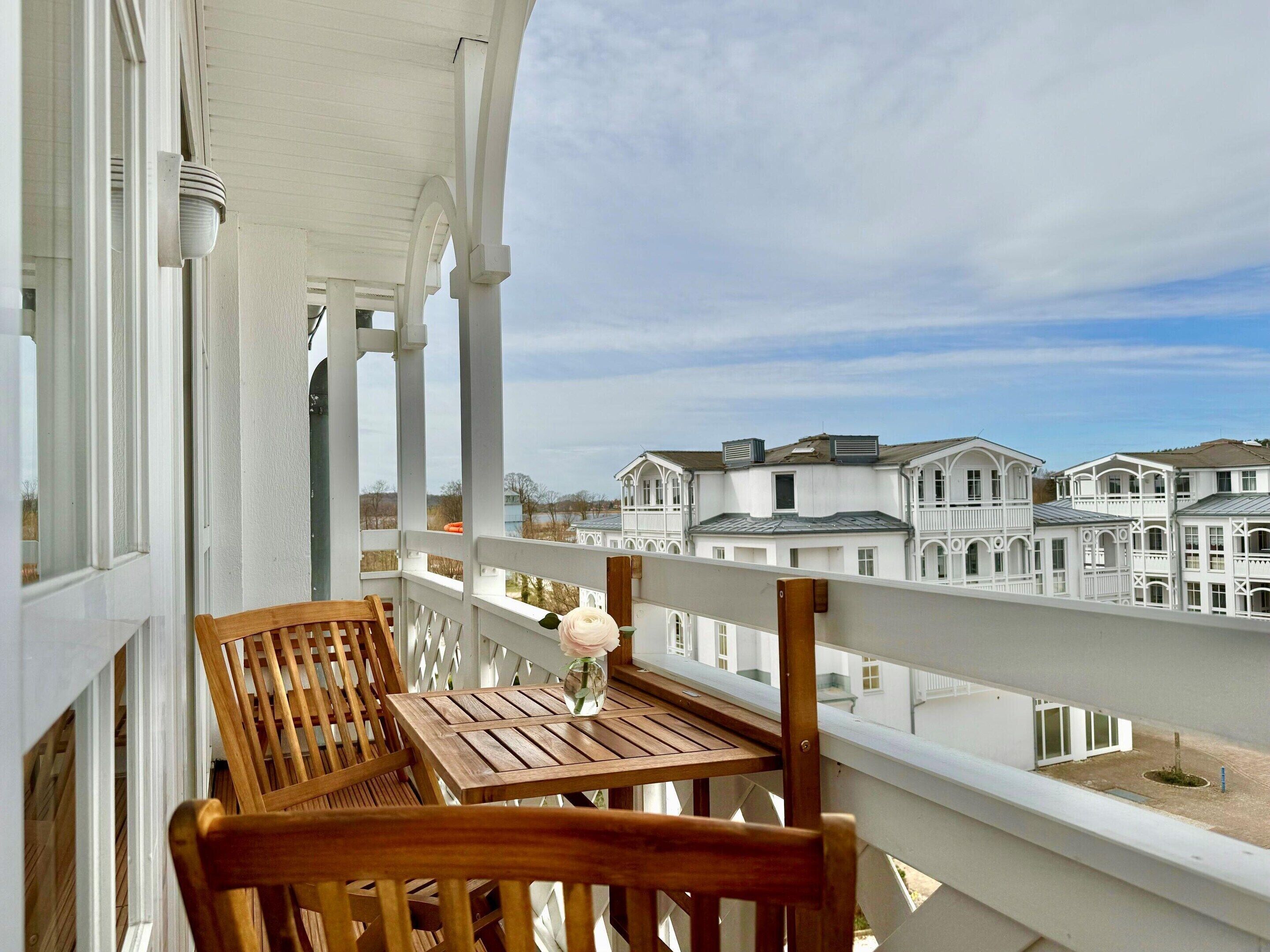A balcony with a wooden table and chairs, overlooking a row of white buildings with balconies.