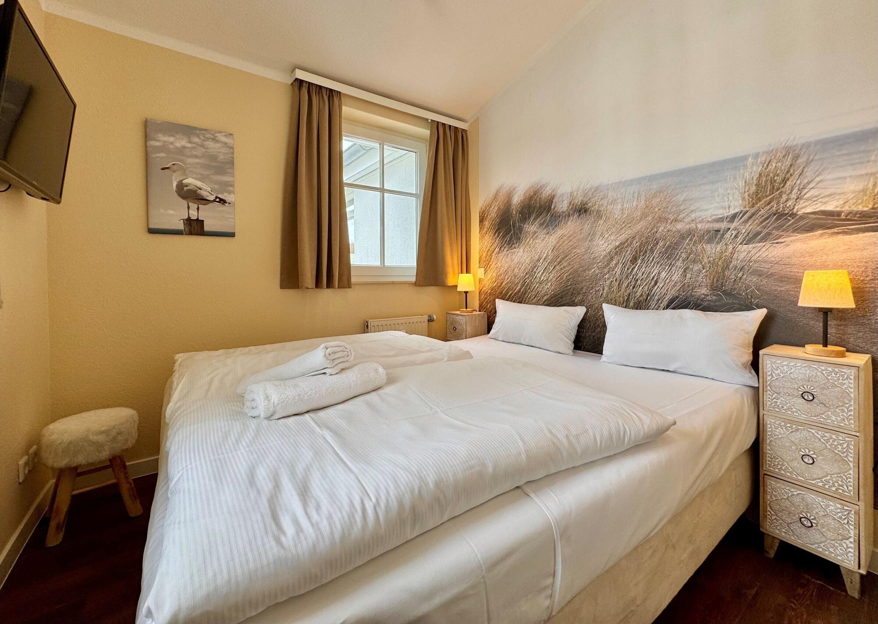 A neatly made bed with white linens and a rolled towel, a bedside lamp, a small stool, and a painting of a seagull on the wall.