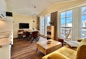 A modern living room with a wooden coffee table, a sofa, a dining area with a TV, and large windows with a view. - Stilvolle Ferienwohnung mit Balkon und Wlan in Sellin (Sellin)