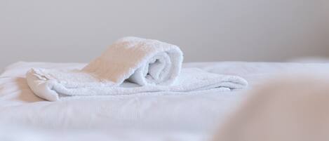 A neatly folded white towel on a bed with a plain white wall in the background.