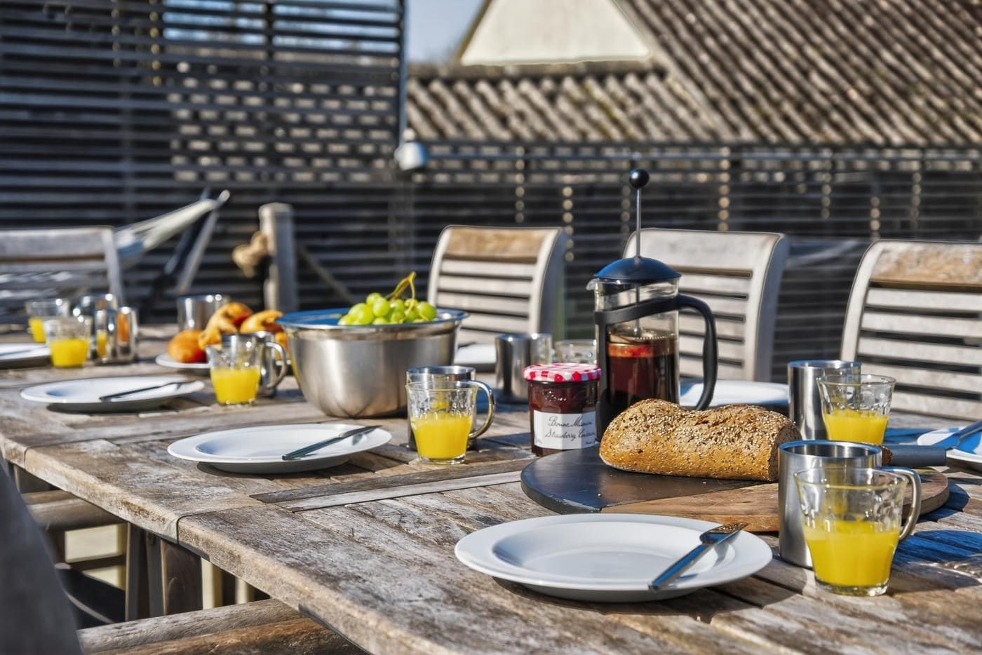 An outdoor dining table set with plates, cutlery, a bread roll, a jug of juice, a pot of coffee, and a bowl of grapes.