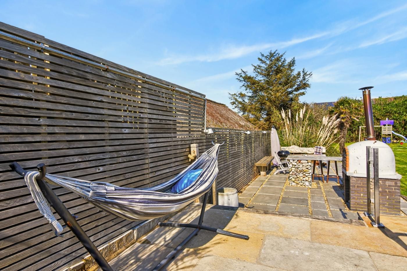 A hammock tied to a wooden fence, a barbecue grill, and a pizza oven in a backyard setting.