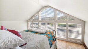 Une chambre avec un plafond en pente, un lit avec des oreillers, une couverture colorée et de grandes fenêtres équipées de stores.