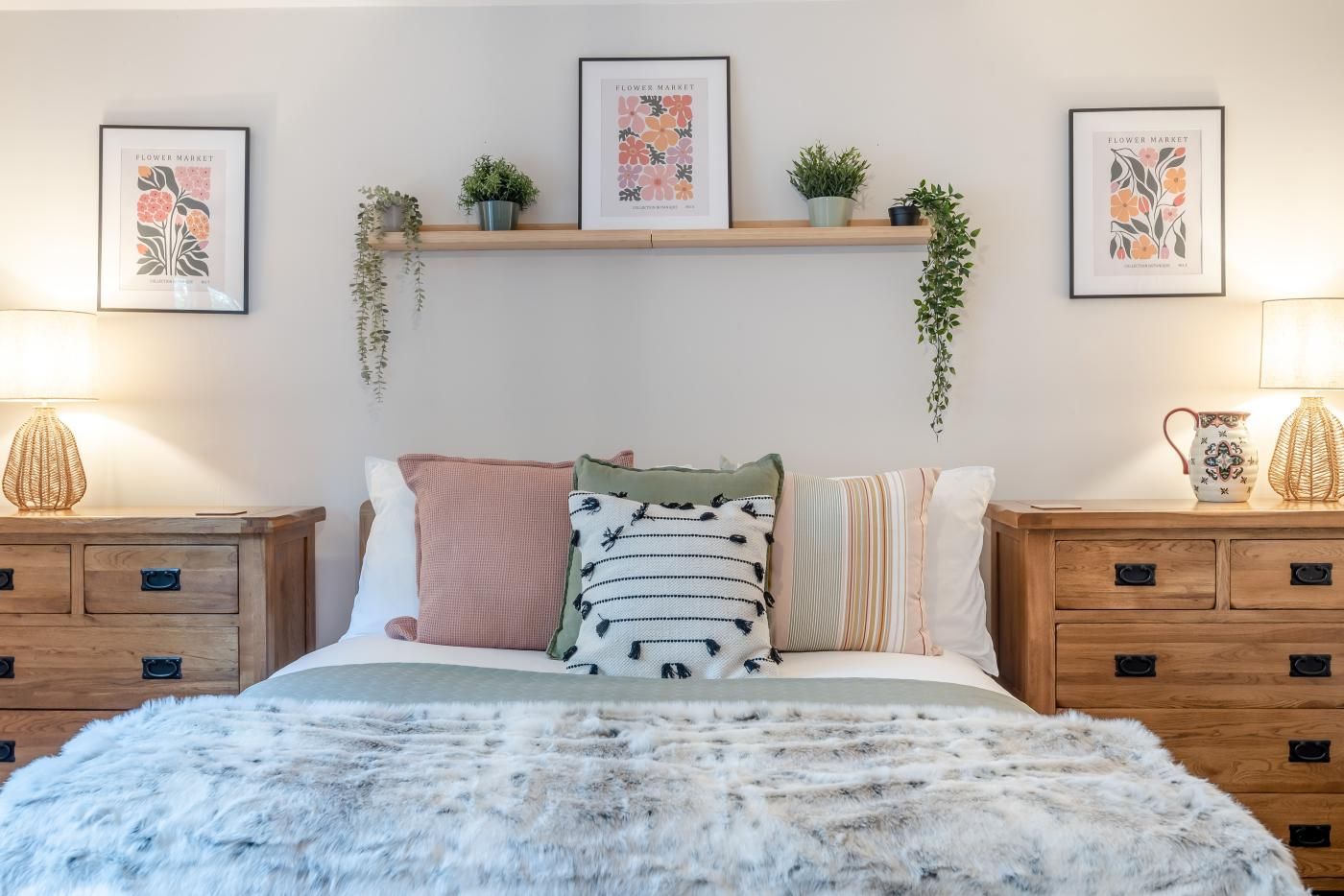 A bedroom with a bed, bedside tables, a shelf with plants, and framed artwork on the wall.